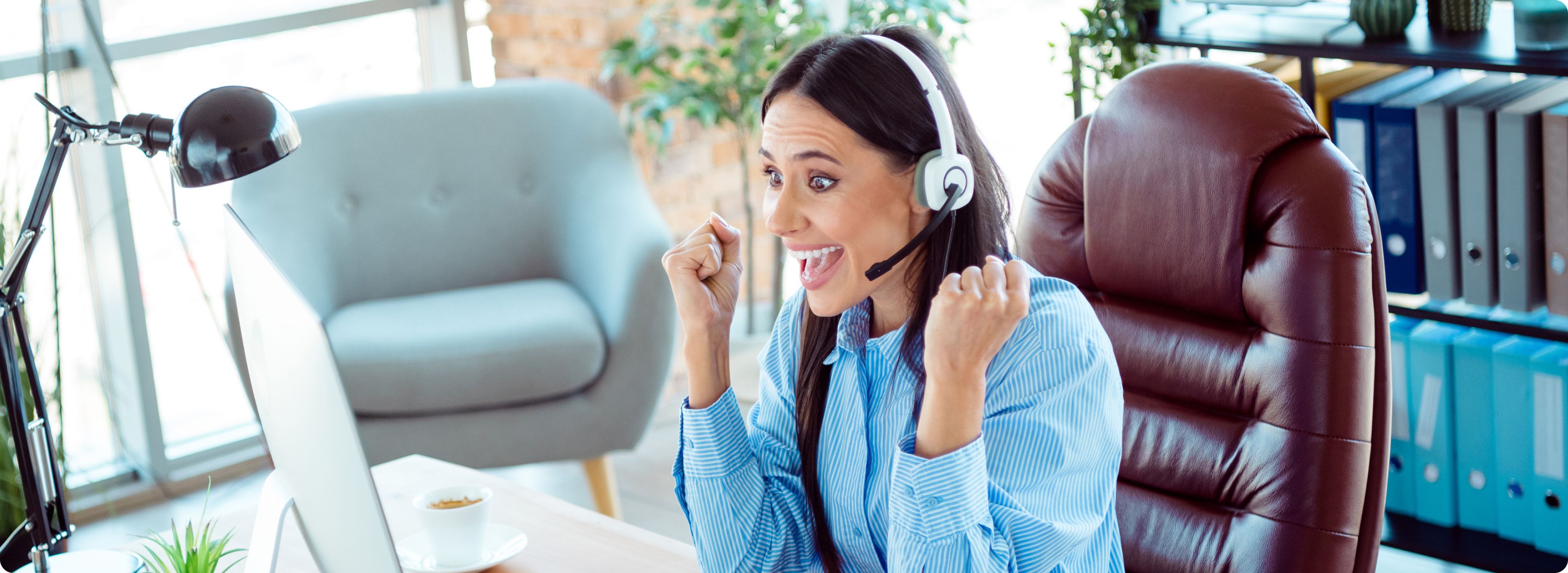 A woman in a blue striped shirt excitedly raises her fists, wearing a headset in front of a computer. Her expression conveys enthusiasm and success.