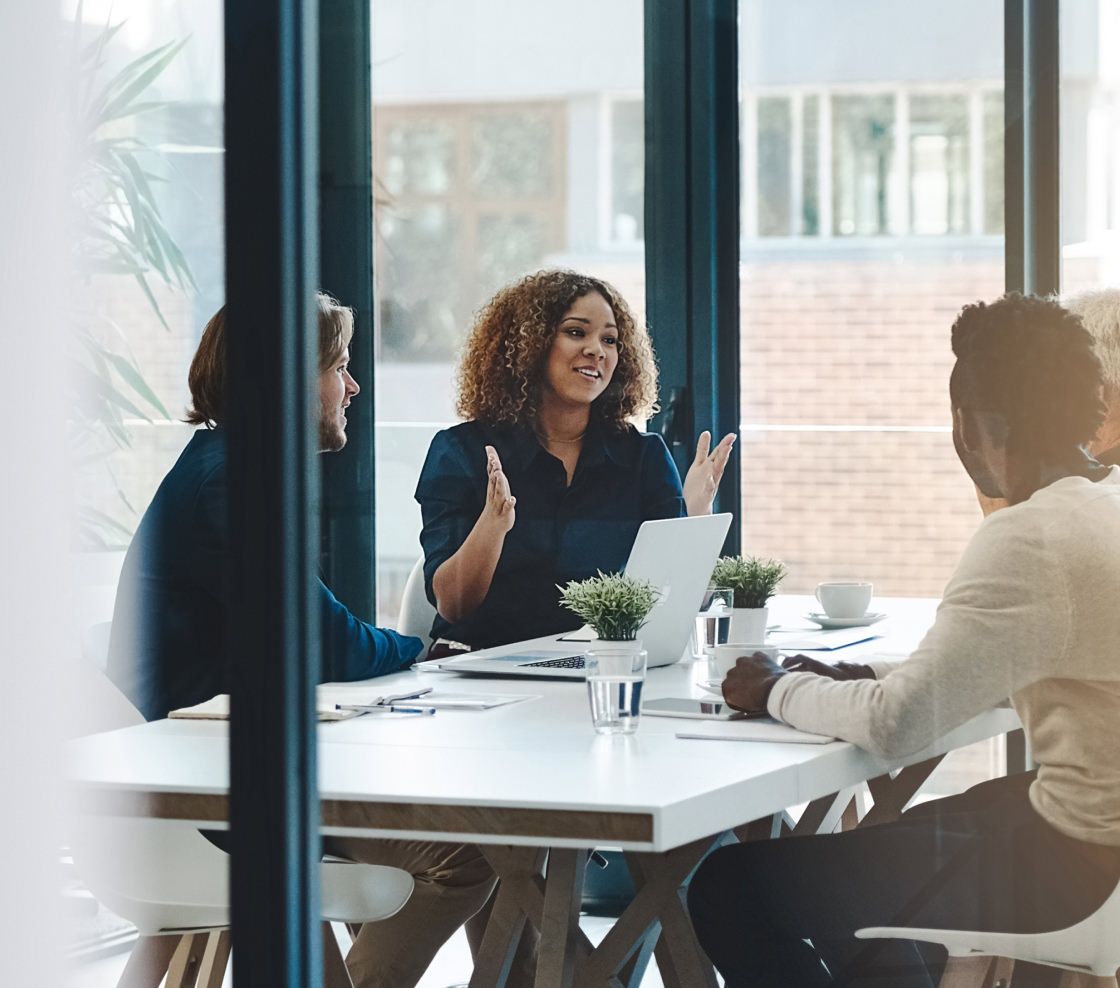 A diverse group of three people are engaged in discussion around a bright office table with laptops and coffee, conveying a collaborative and positive atmosphere.