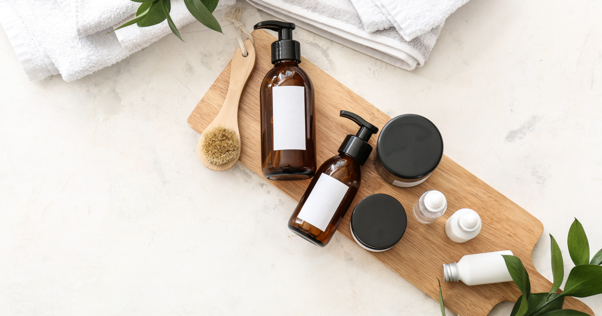 Skincare products grouped as a set on a wooden board, including amber pump bottles, small jars, and dropper bottles, with white towels and green leaves on a marble surface