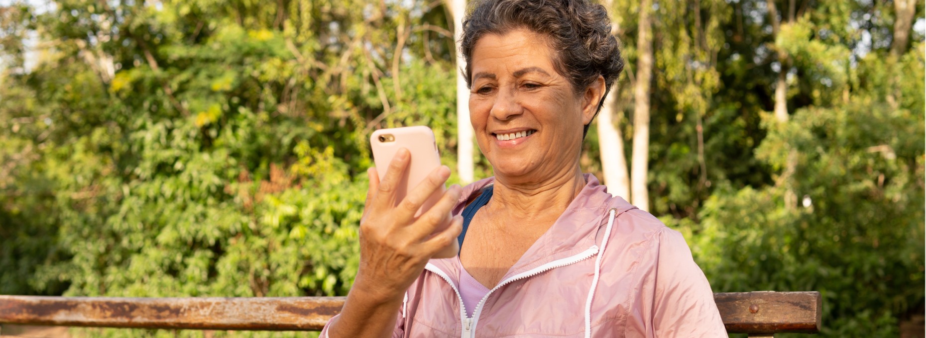 Smiling woman in a pink windbreaker on a park bench, reading a reply from a brand on her phone