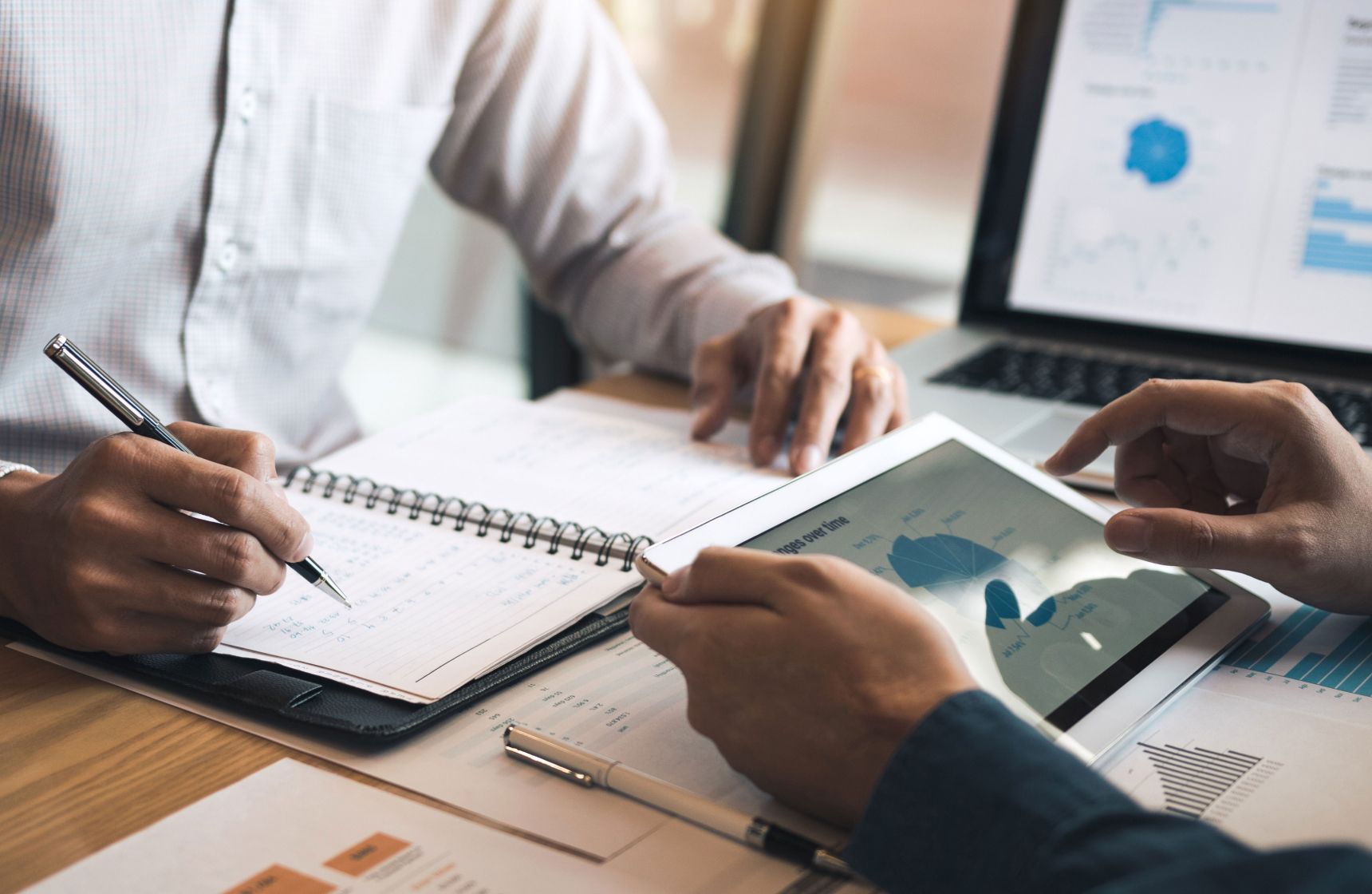 An image showing hands writing in a notebook and using an ipad for work