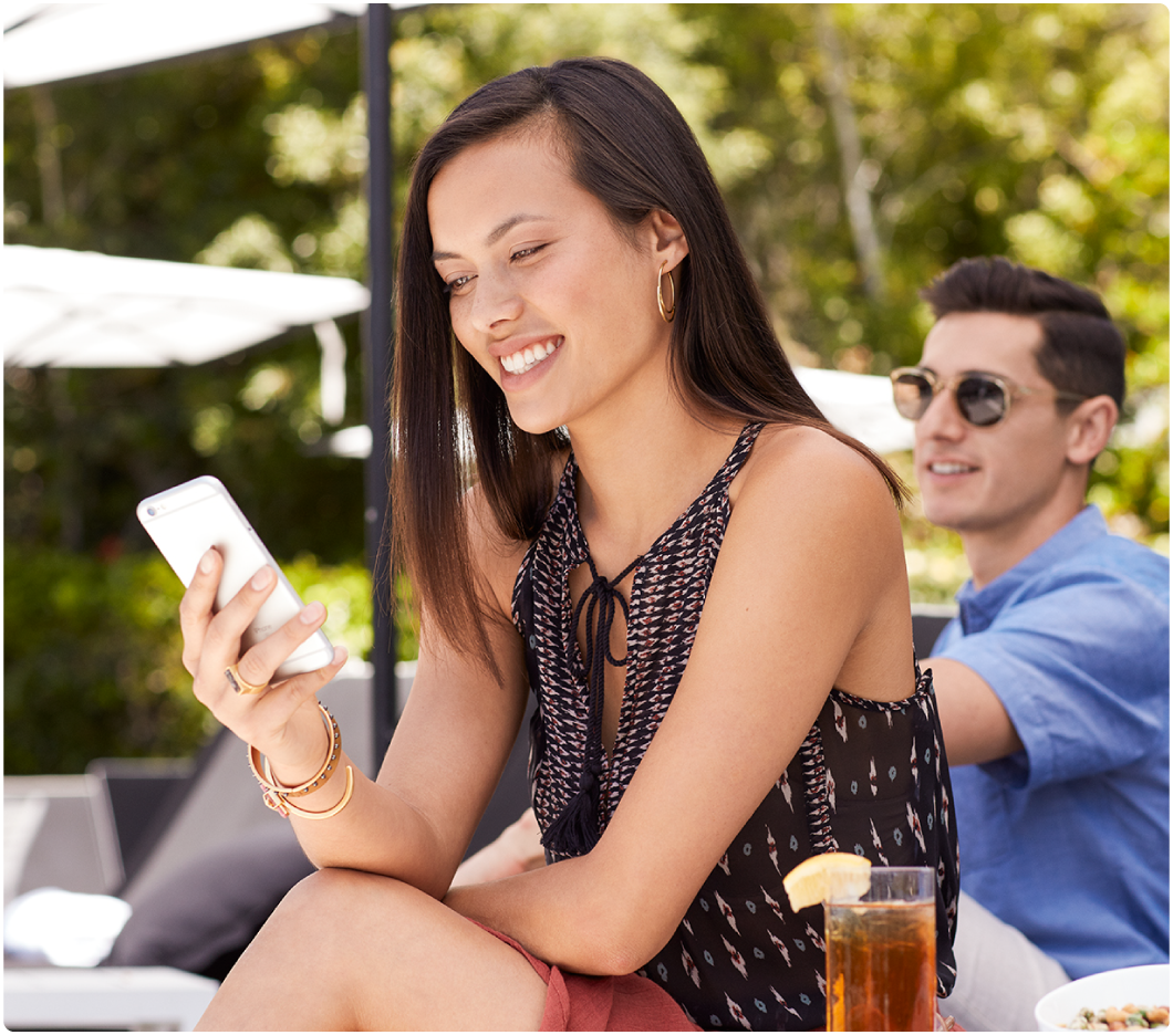 A woman smiling at her phone sits outdoors, with a man in the background wearing sunglasses. A glass of iced tea is on the table. Relaxed and sunny atmosphere.