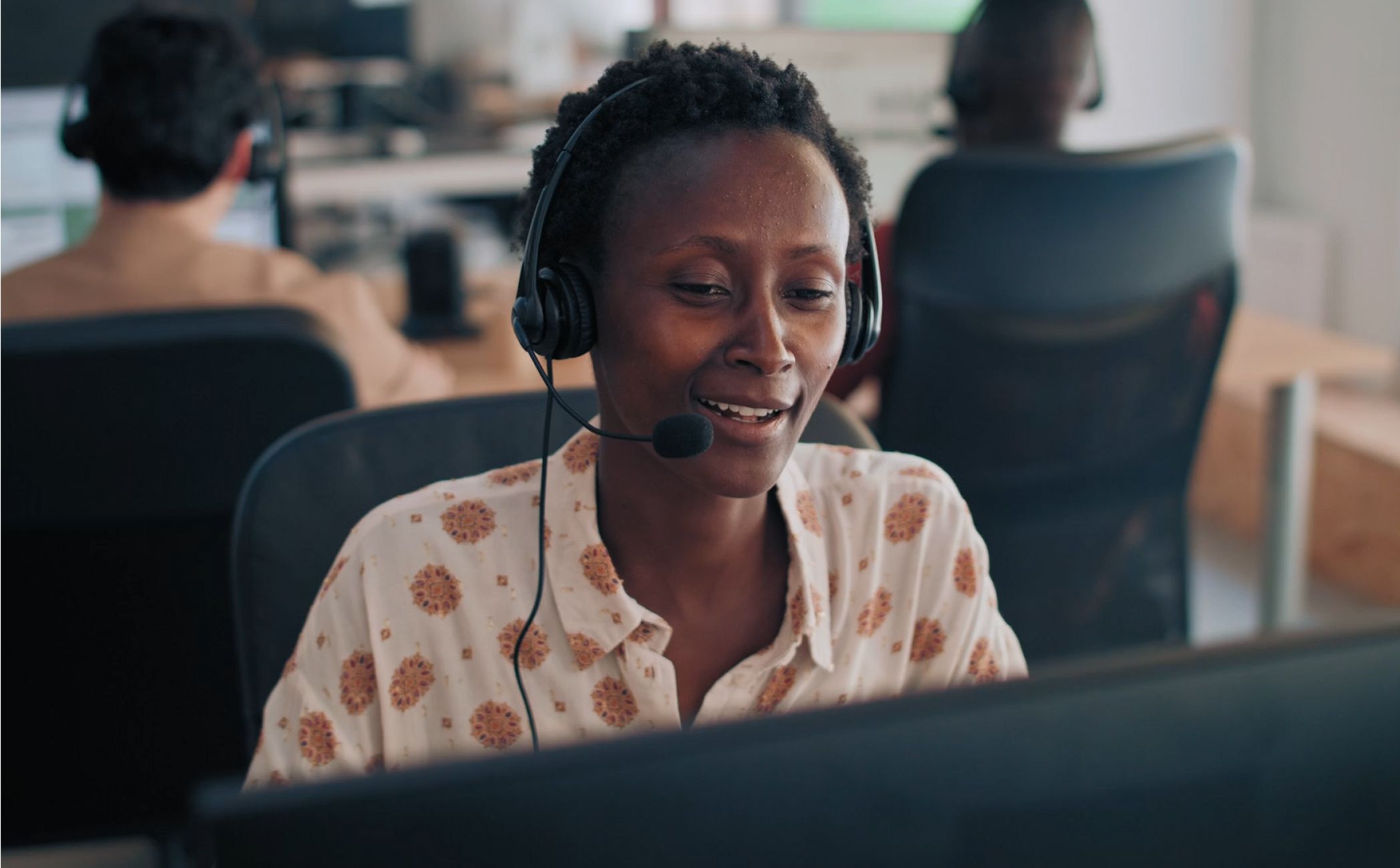 A woman wearing a patterned blouse and headset smiles while working at a computer in a lively office environment, conveying a cheerful, professional tone.