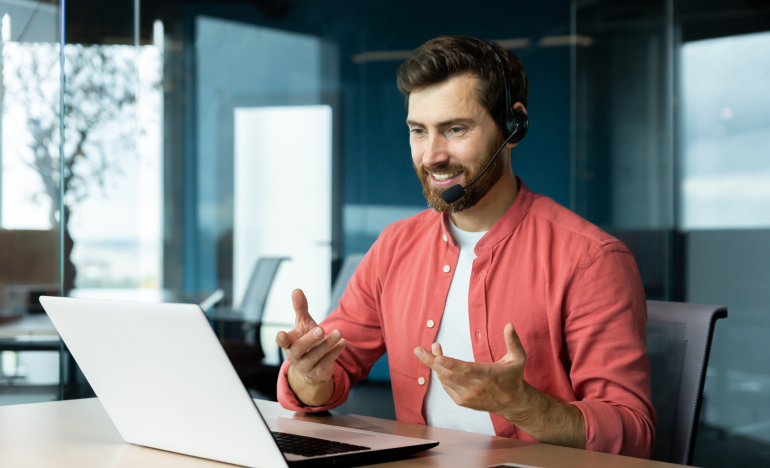 Bearded man wearing a phone headset and a salmon-colored shirt gesturing at a laptop