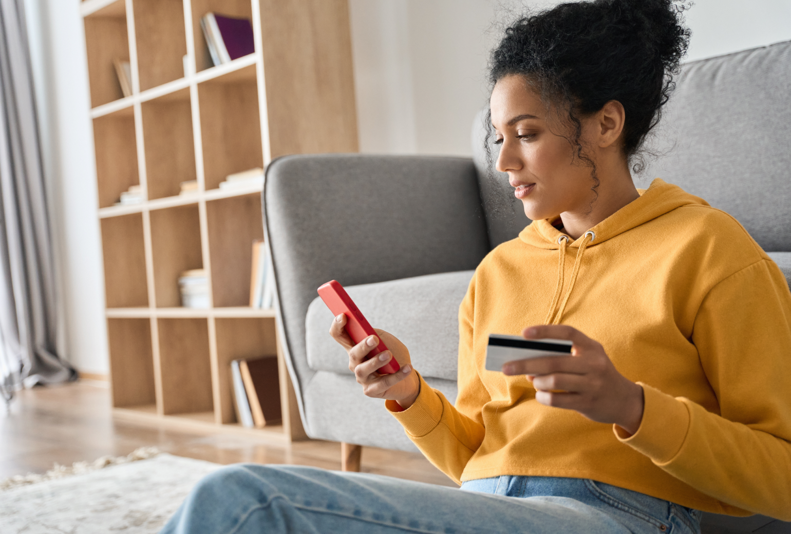 A woman in a yellow sweatshirt in her living room buying something online with her red smartphone, a credit card in her hand