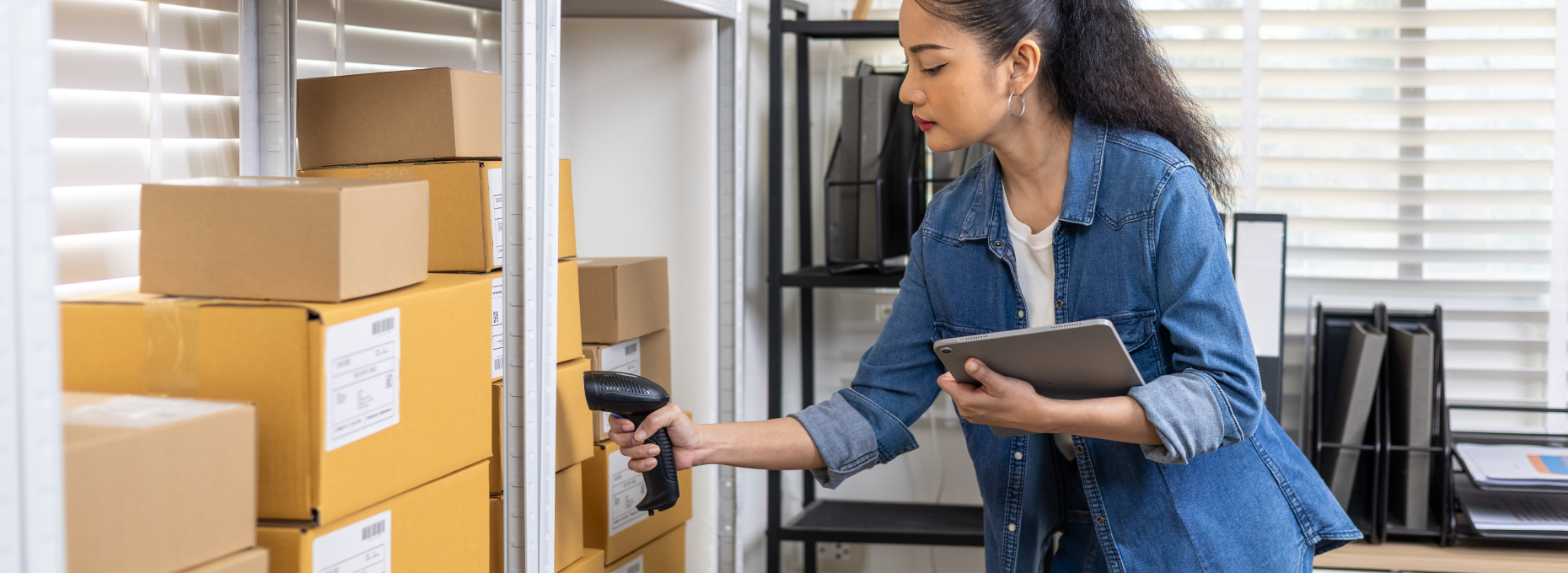 A small business owner scans a product with a handheld barcode reader while holding a tablet, standing at a shelving unit stocked with boxes near a sunlit window.