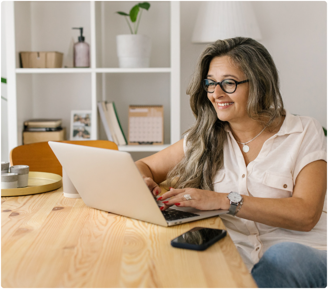 A smiling woman with long hair and glasses works on a laptop at a wooden desk. The background features shelves with plants and books, creating a cozy ambiance.