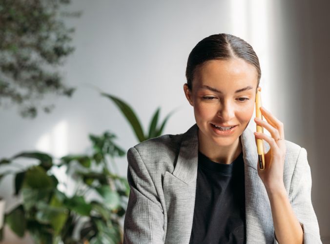 A woman in a gray blazer smiles while talking on a smartphone, seated indoors with green plants in the background. The atmosphere is professional and friendly.