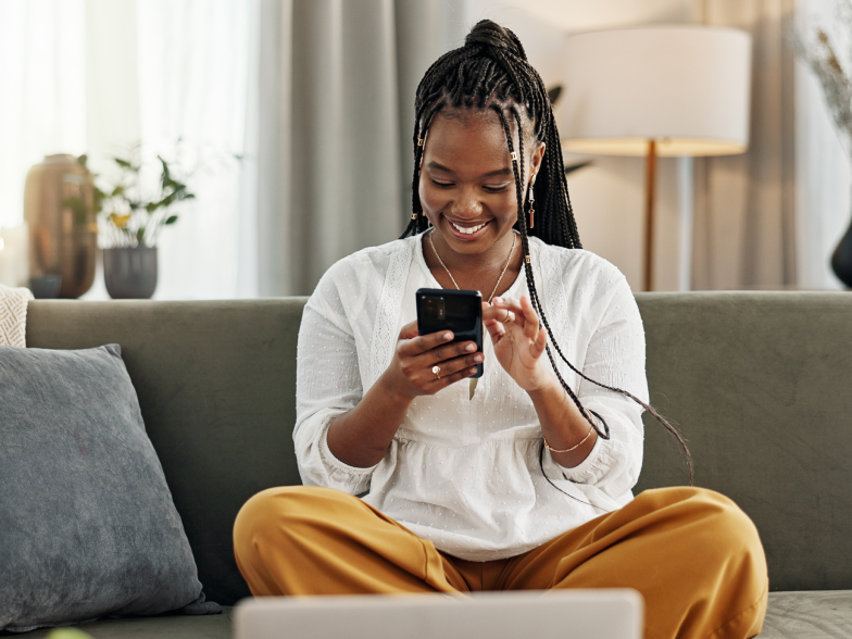 Woman sitting cross-legged on a gray couch at home, smiling as she shops on her phone