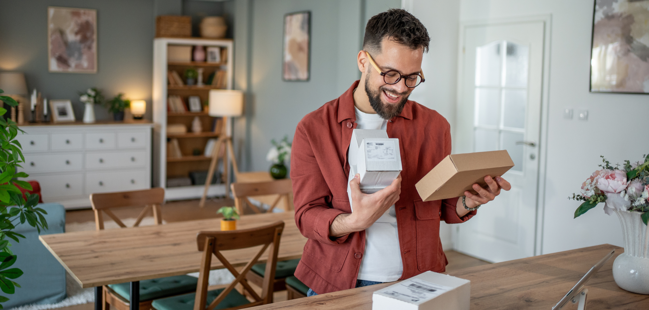 A man with glasses smiles while holding a small package in a cozy, well-lit living room. The room features wooden furniture and decorative plants, creating a warm atmosphere.