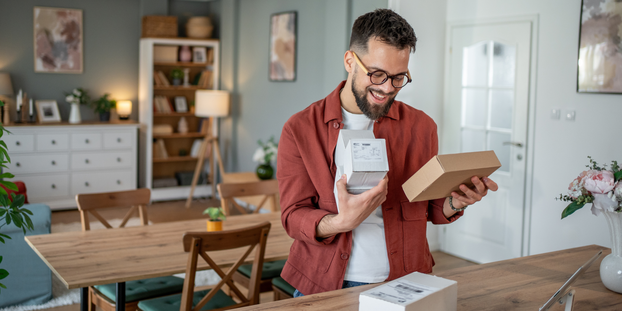 A man with glasses smiles while holding a small package in a cozy, well-lit living room. The room features wooden furniture and decorative plants, creating a warm atmosphere.