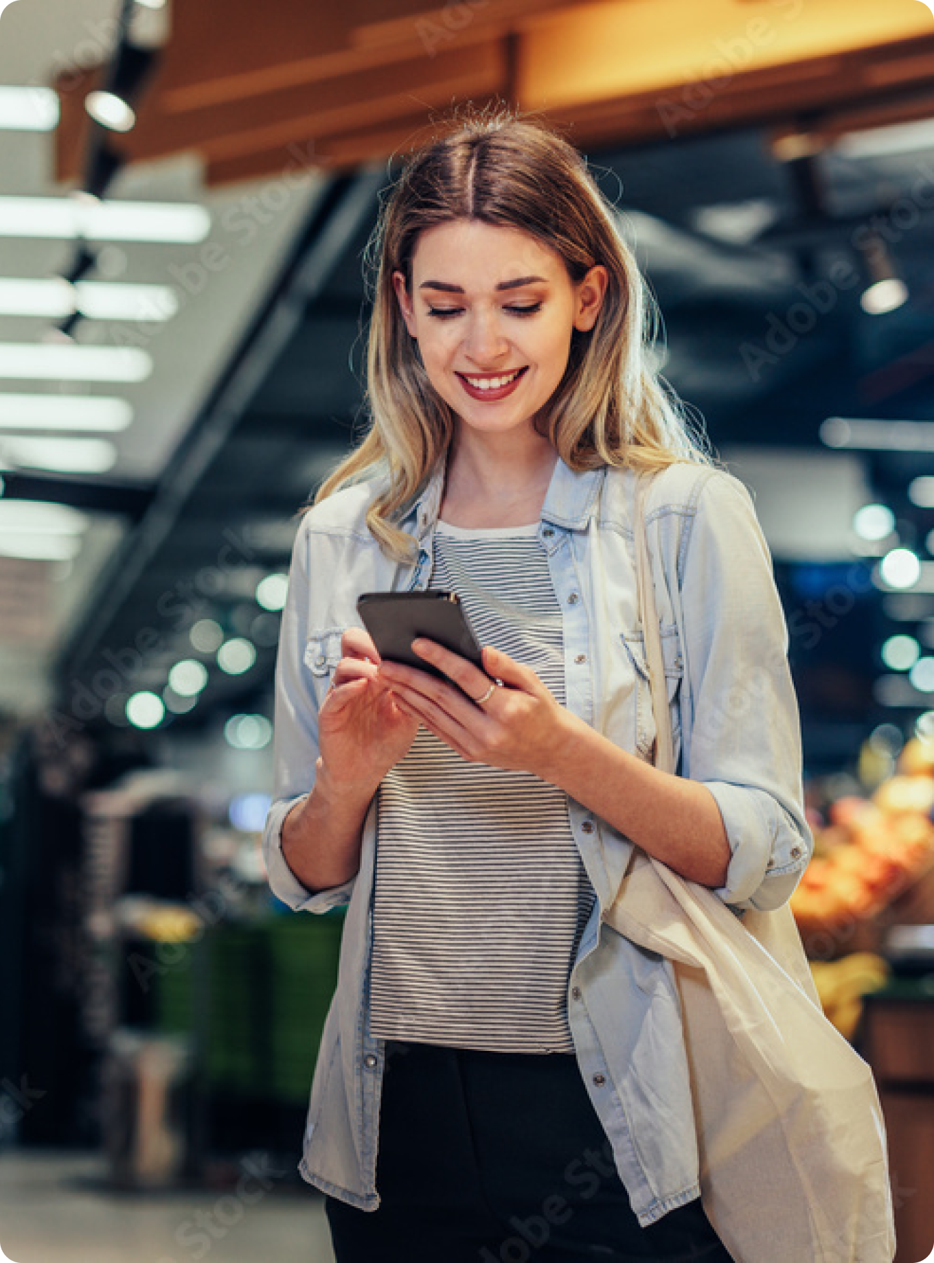 Young woman in a light denim shirt smiles while looking at her phone in a supermarket aisle. She carries a reusable bag, conveying a casual tone.
