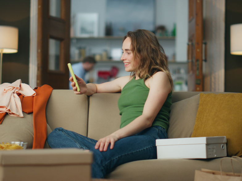 A woman in a green tank top sits on a living room couch in the evening, smiling at her phone. Lamps light the room and a few clothes are draped over the arm of the couch.