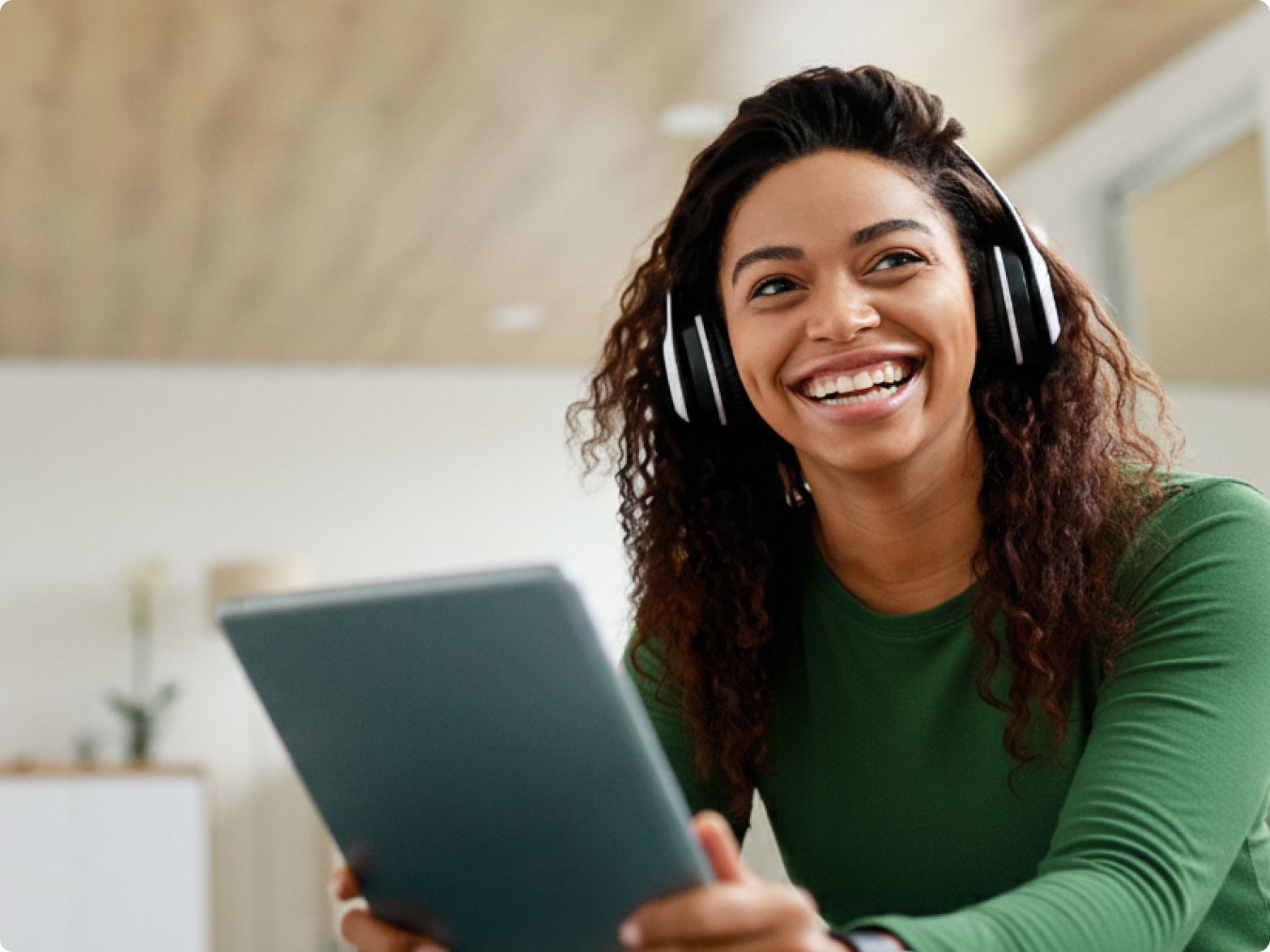 Smiling woman wearing headphones and a green shirt looks to the side while holding a laptop. She is seated in a bright room, creating a joyful atmosphere.