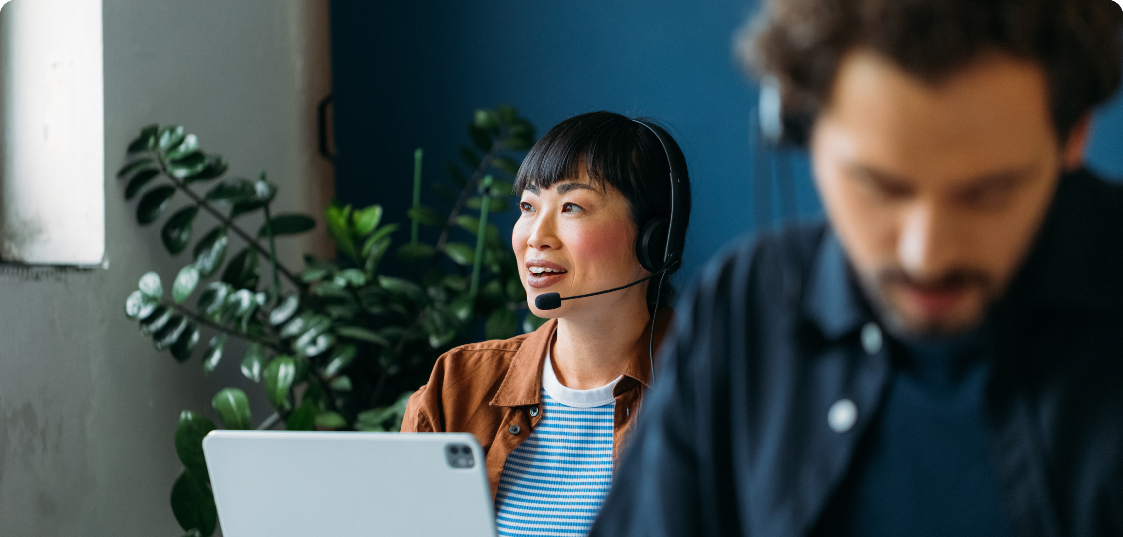 A woman with a headset smiles, engaged in a video call at a laptop in a bright office with plants. A blurred colleague works in the foreground.