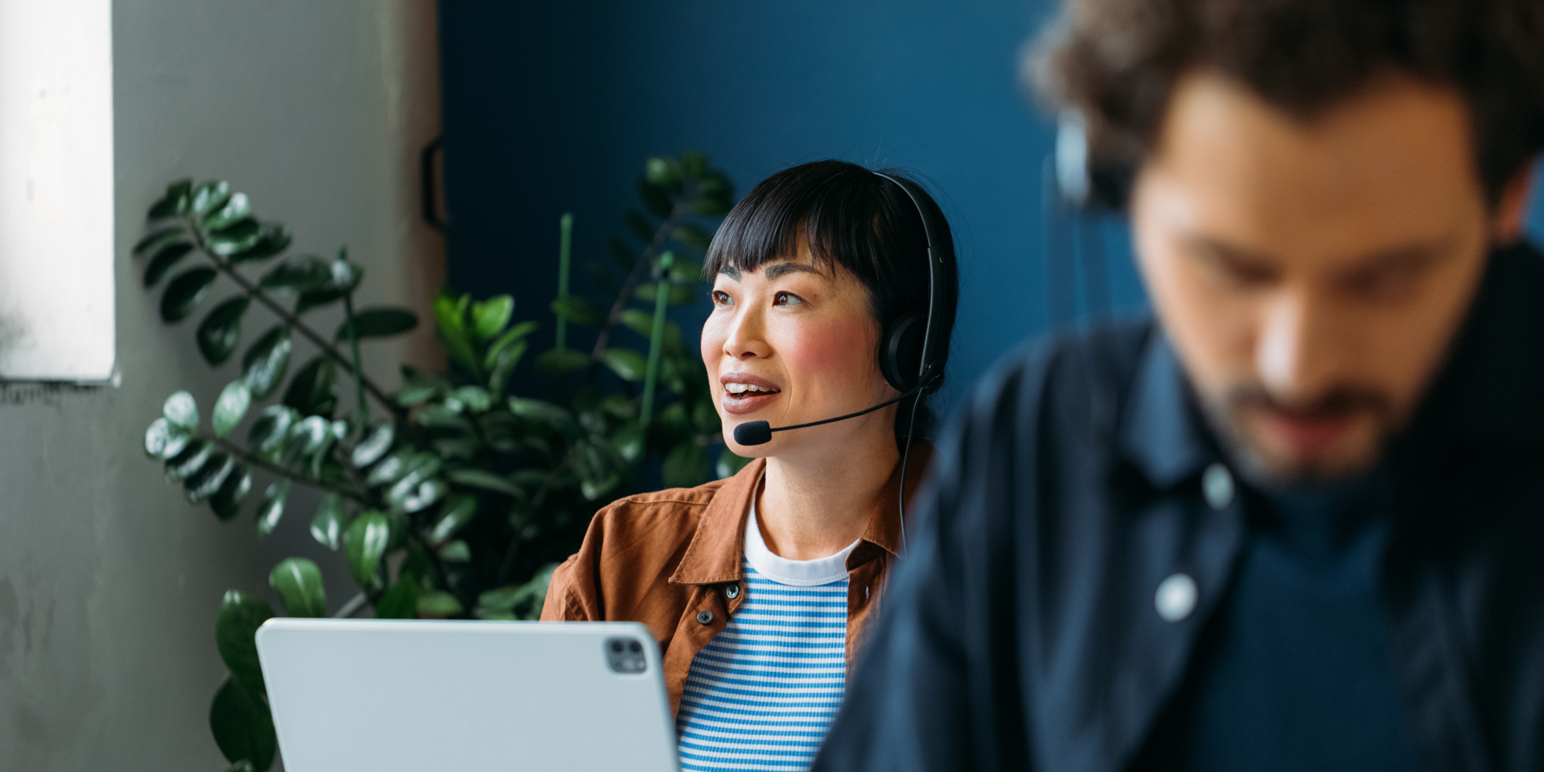 A woman with a headset smiles, engaged in a video call at a laptop in a bright office with plants. A blurred colleague works in the foreground.