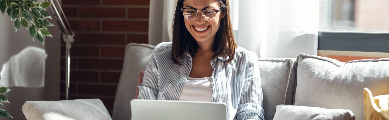Woman smiling at the laptop in her lap while seated on a gray couch