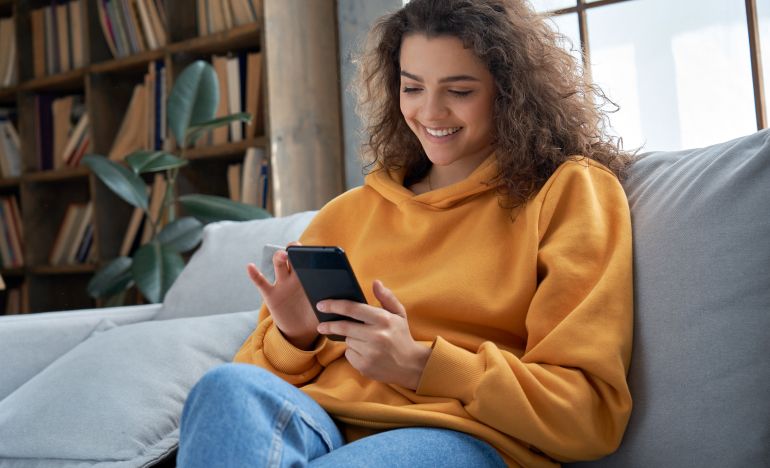 A woman in an orange sweatshirt sitting in her living room on her smartphone