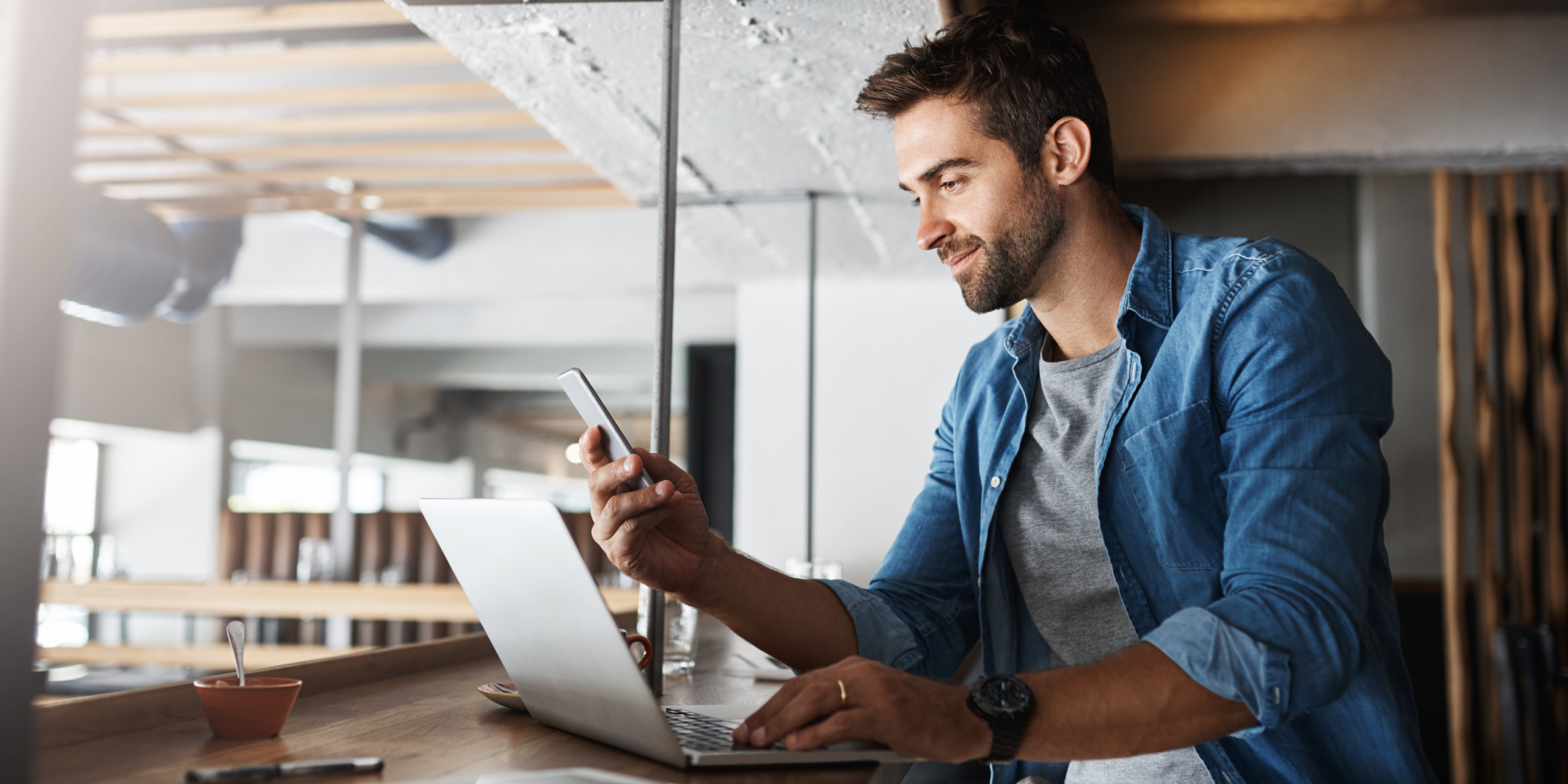 Man in a casual blue shirt smiles while multitasking with a laptop and smartphone at a stylish, modern cafe, conveying productivity and ease.