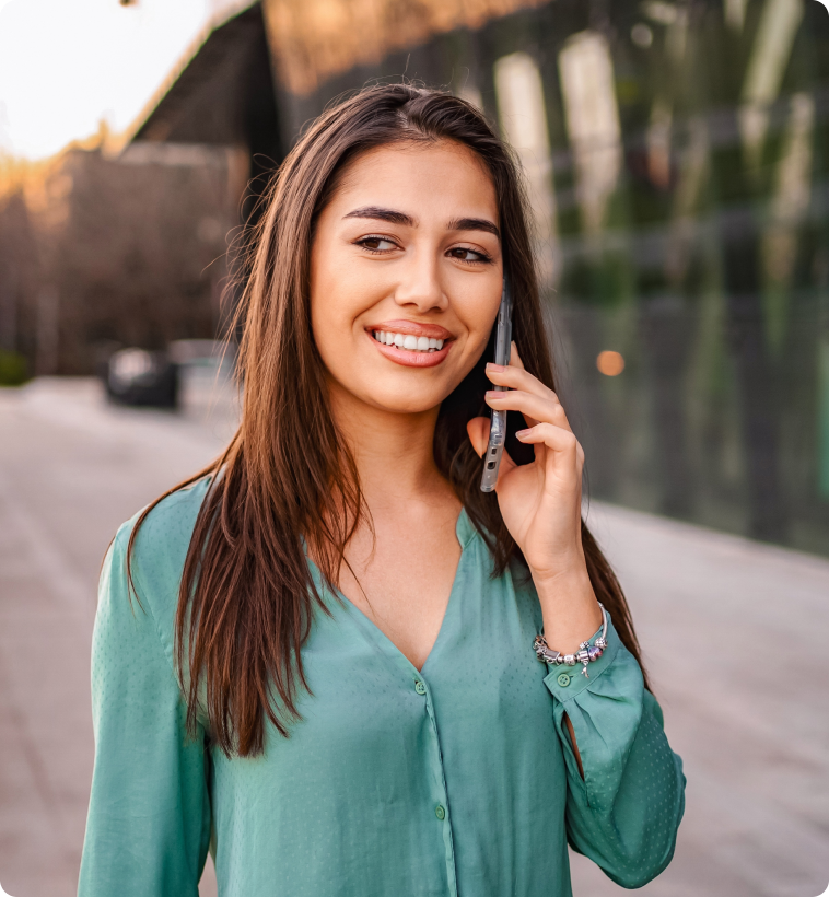 Smiling woman in a green blouse talks on her phone while walking outside. Background shows a blurred urban setting with soft evening light.