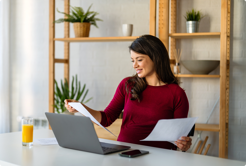 A woman in a red shirt smiles while holding documents in front of a laptop. A glass of orange juice and a phone rest on the table, with plants on shelves behind her.
