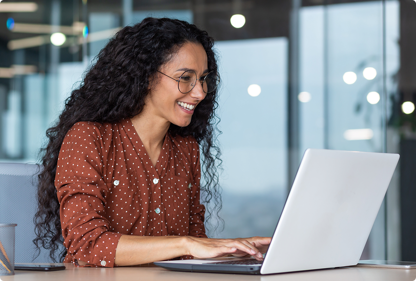 Smiling woman with curly hair and glasses types on a laptop in a modern office. She wears a brown polka dot blouse, conveying a cheerful mood.