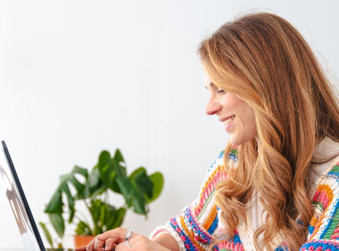 A woman with long hair smiles while typing on a laptop. She's wearing a colorful sweater. A green potted plant is blurred in the background.
