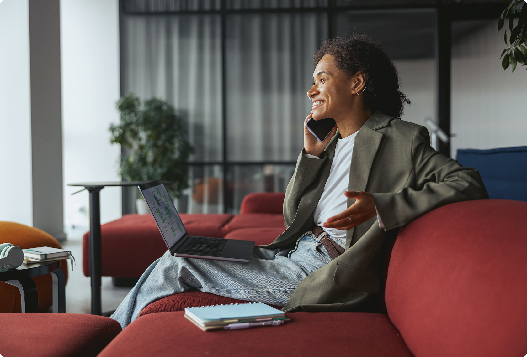 Smiling woman talking on phone, sitting on a red sofa with a laptop on her lap. Casual setting with a relaxed, professional atmosphere.