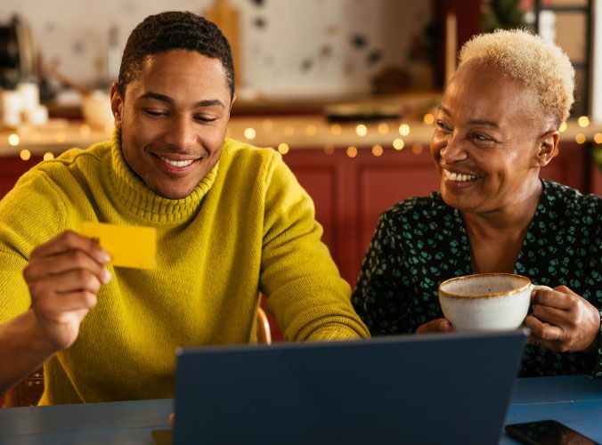 Smiling man next to his mother, holding a credit card to buy something on laptop