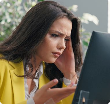 A woman in a yellow blazer looks frustrated while staring at a computer screen, resting her head on her hand. Plants are blurred in the background.