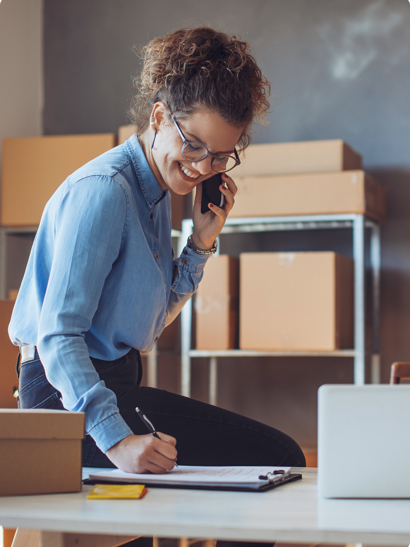 A woman in a denim shirt talks on the phone while writing on a clipboard in a room with cardboard boxes, conveying a busy and organized atmosphere.