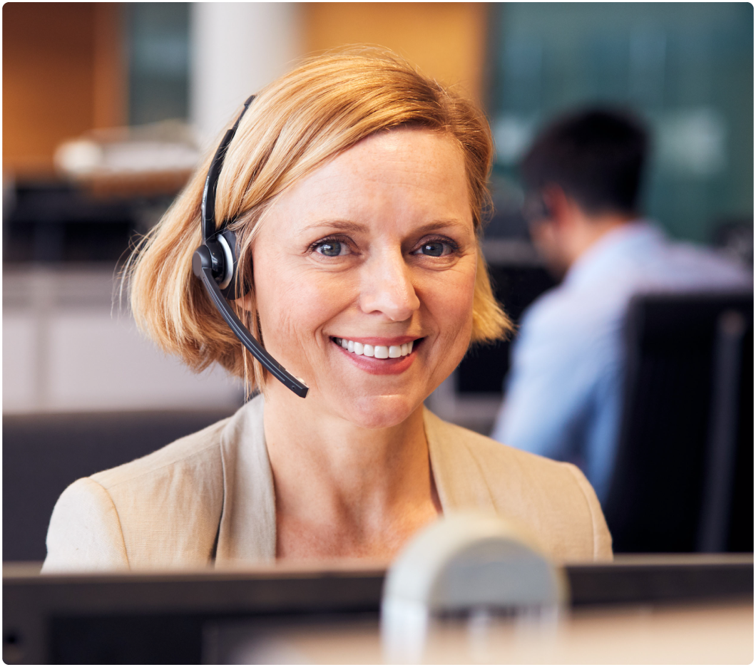A smiling woman wearing a headset in an office setting, foregrounded against a blurred background with a colleague seated at a desk. She appears engaged and approachable.