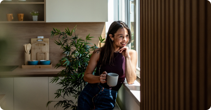 Smiling woman in a kitchen leaning on a windowsill, holding a mug. Sunlight filters in, illuminating her face. Warm, relaxed atmosphere.
