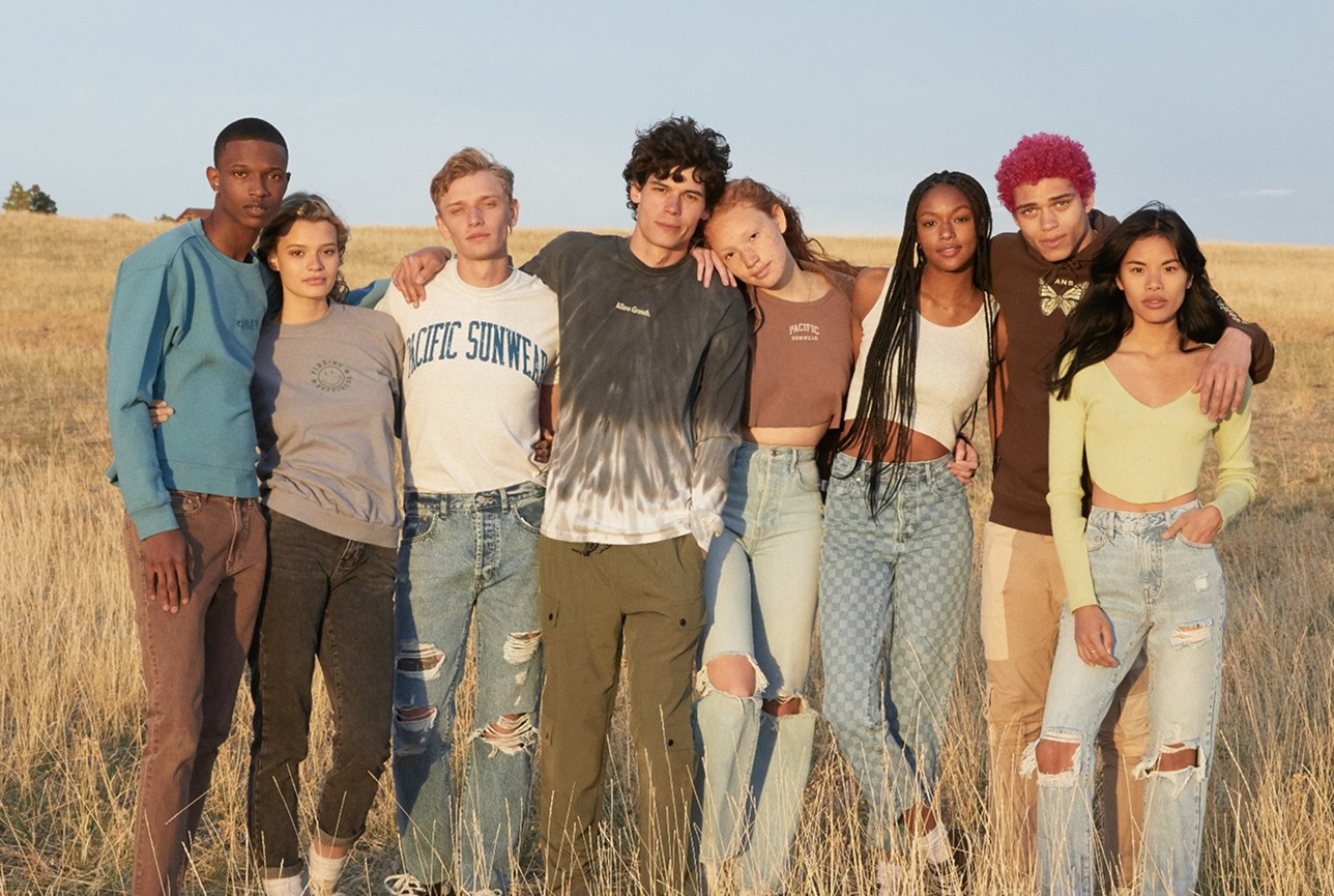 A diverse group of eight young adults stands closely in a sunlit field, smiling warmly. They wear casual, trendy outfits, conveying a relaxed, friendly vibe.