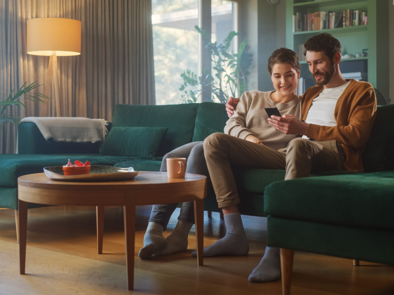 Couple relaxing on a green sofa in a sunlit living room, shopping together on a smartphone