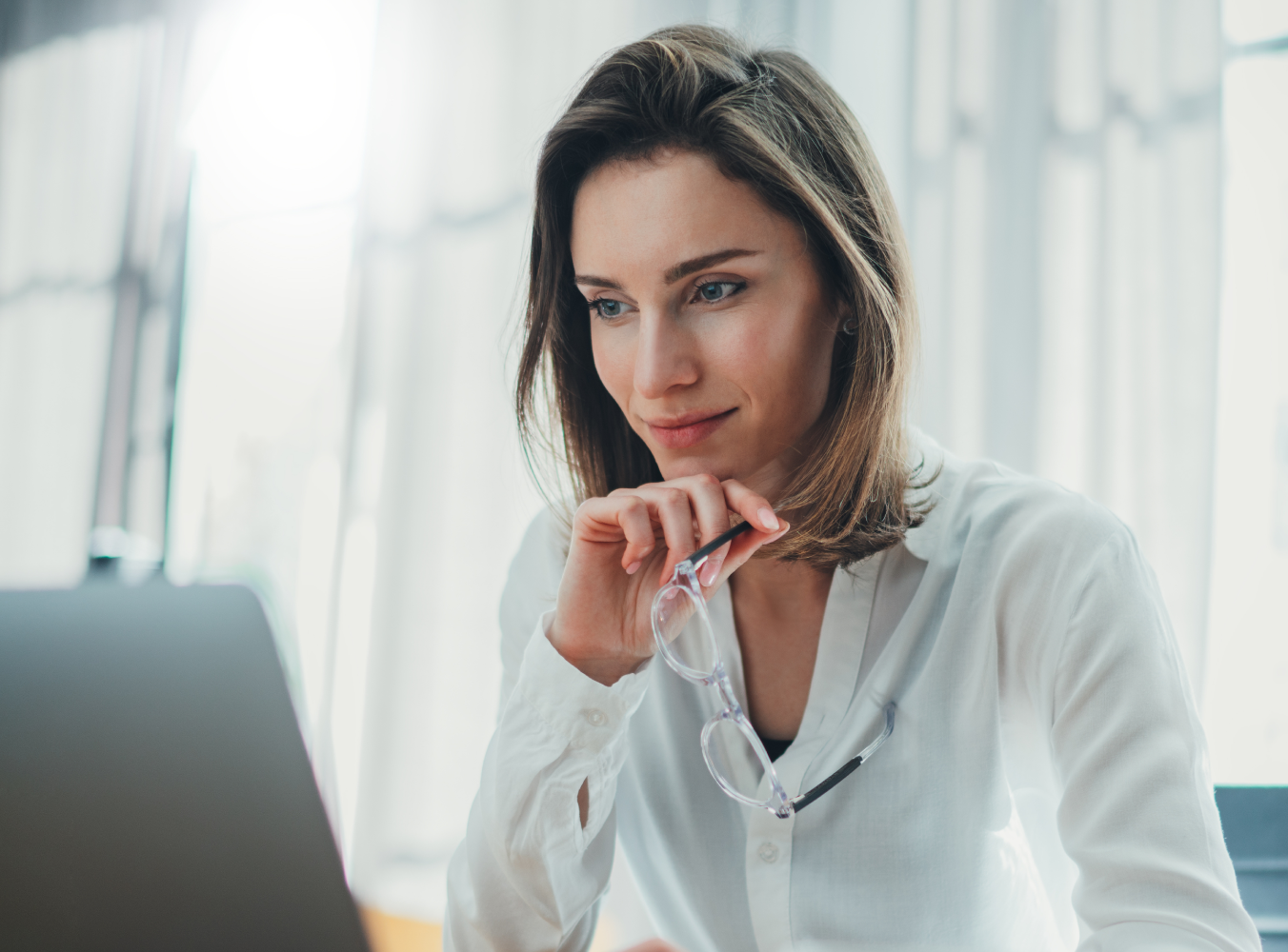 A woman in a white blouse sits at a desk, gazing thoughtfully at a laptop while holding eyeglasses. The scene is bright and focused, suggesting concentration.