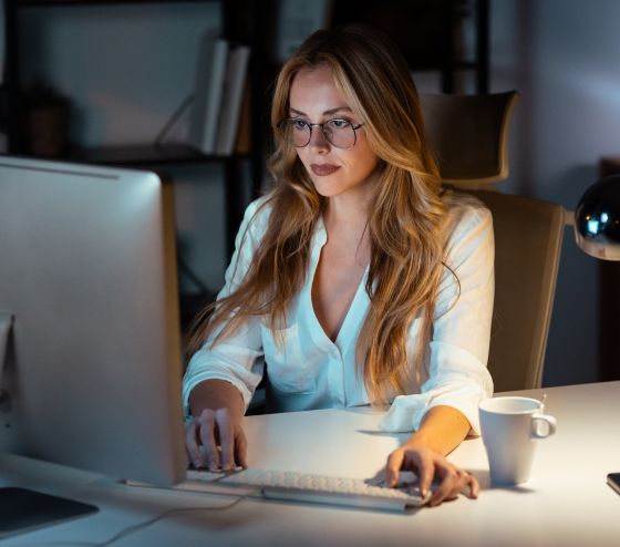 A woman with long hair and glasses is focused on a computer in a dimly lit office. A cup sits nearby on the desk, conveying a quiet, studious atmosphere.