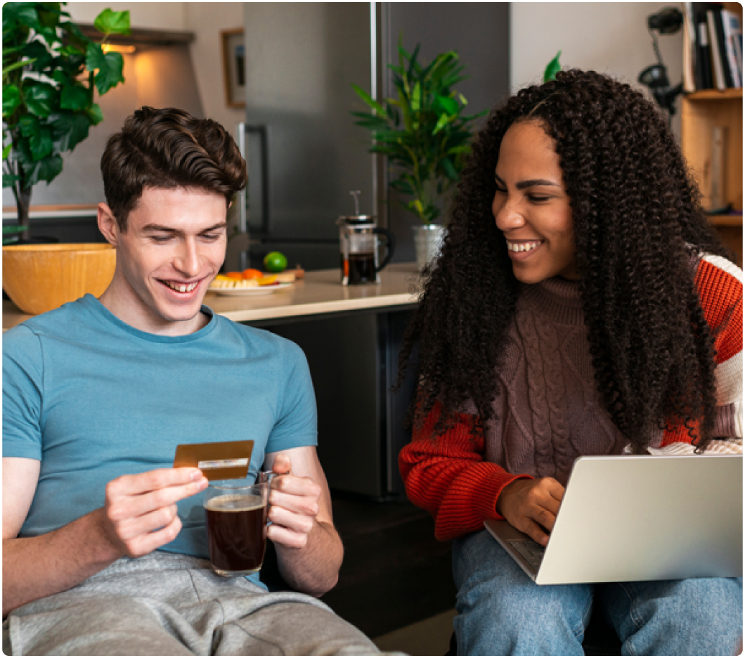 A man in a blue shirt, holding a credit card and coffee, smiles while a woman with curly hair and a laptop laughs next to him in a cozy kitchen.