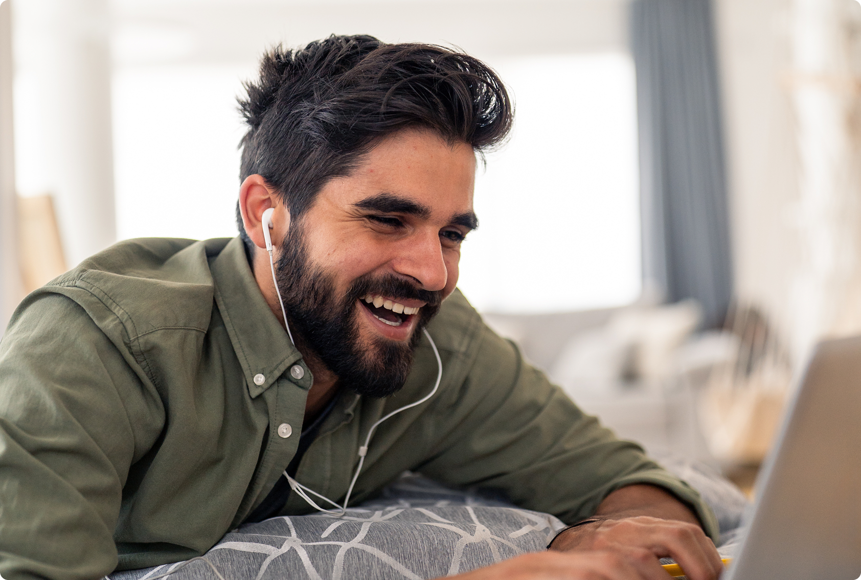 A man with dark hair and a beard, wearing earphones and a green shirt, smiles joyfully while looking at a laptop in a cozy, softly-lit room.