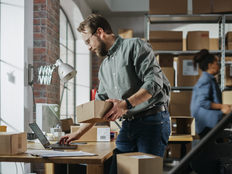 A small business owner checks his laptop while holding a package in a warehouse fulfillment area, with a colleague packing boxes in the background.