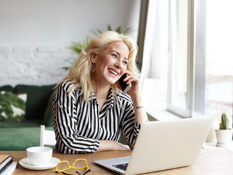 A woman with long blonde hair, wearing a black-and-white striped blouse, laughs while talking on her phone and sitting at a laptop near a bright window.