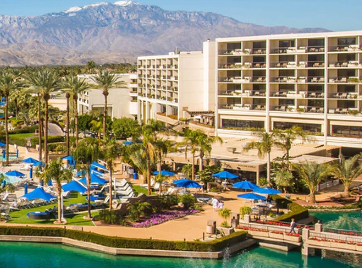 A luxurious resort with tall palm trees, a pool with blue umbrellas, and a modern hotel building. Snow-capped mountains are seen in the background.