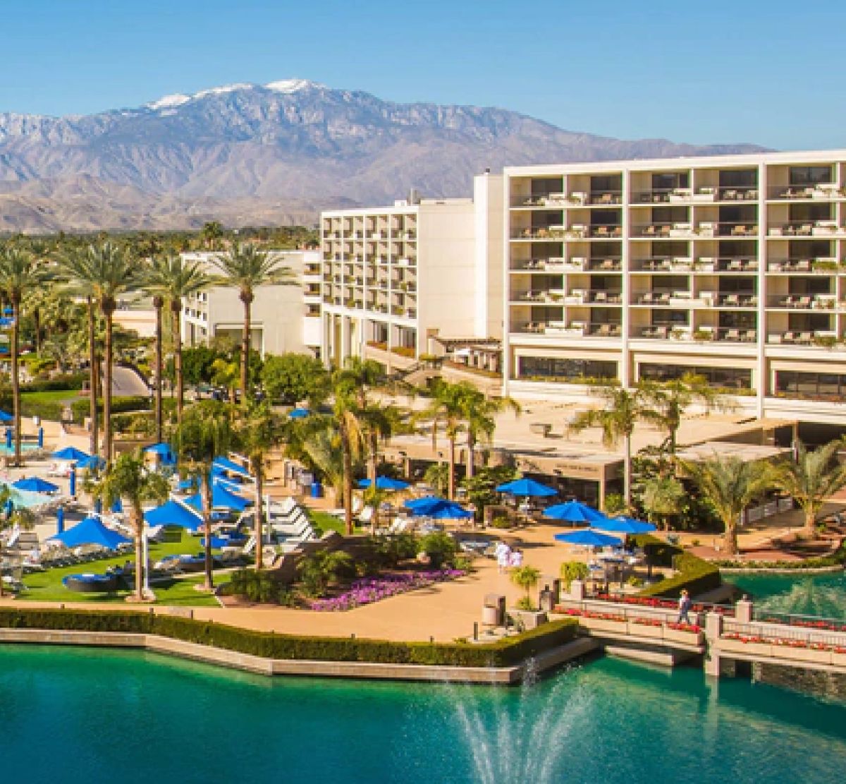 A luxurious resort with tall palm trees, a pool with blue umbrellas, and a modern hotel building. Snow-capped mountains are seen in the background.
