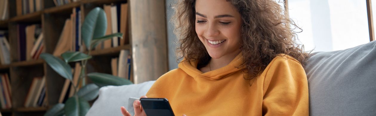 A woman in an orange sweatshirt sitting in her living room on her smartphone