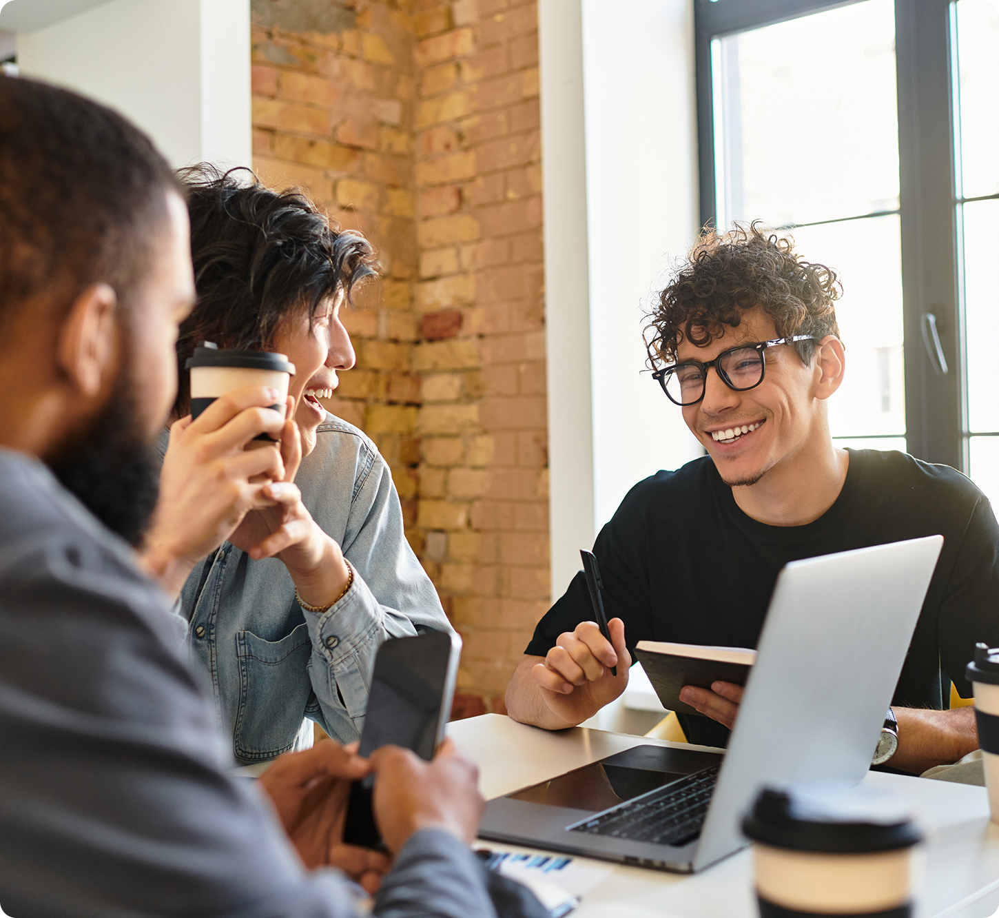 Three people sit around a table in a bright room with brick walls. They are smiling and chatting, holding coffee cups and notebooks, conveying a collaborative and friendly atmosphere.
