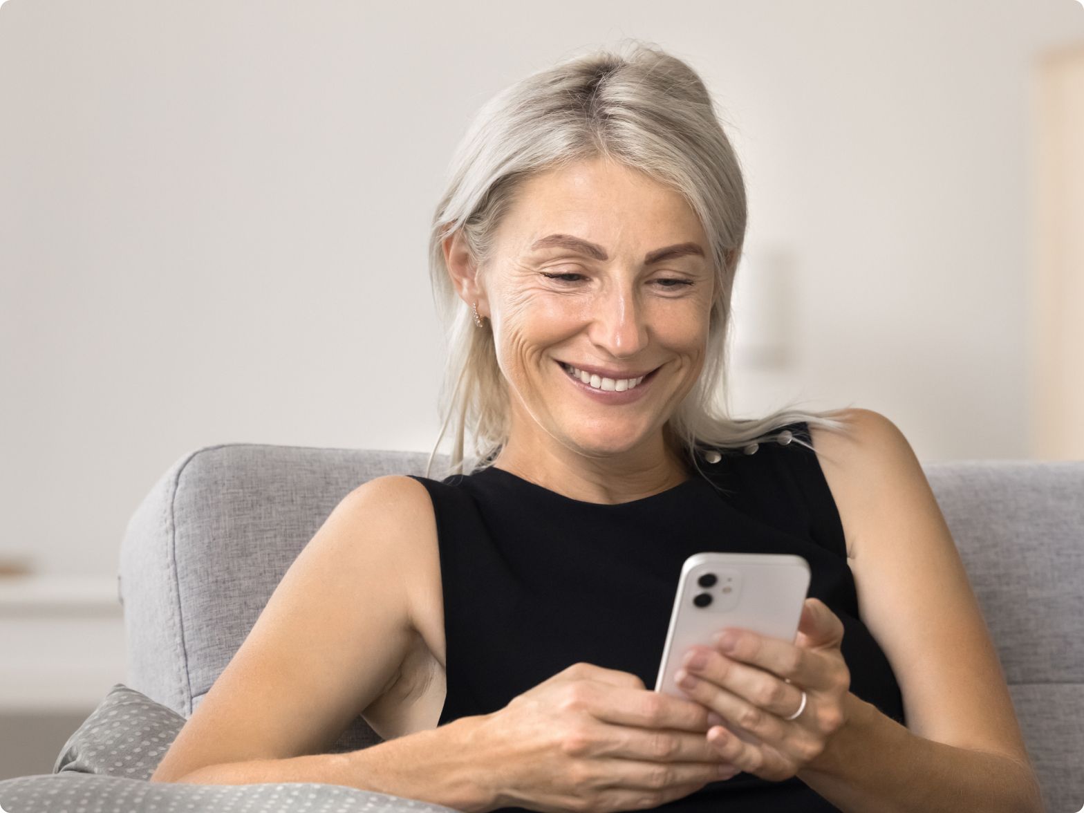 A woman with gray hair smiles while sitting on a couch, looking at her smartphone. She wears a black sleeveless top in a bright, cozy room.