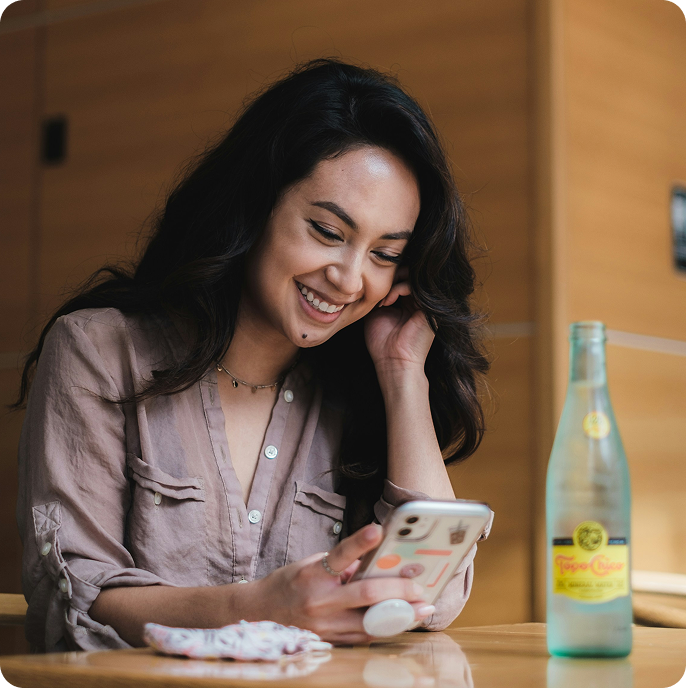 Woman smiling at phone in a cafe, engaged in a chat about exchanging a jacket. Texts on screen express friendly customer service.
