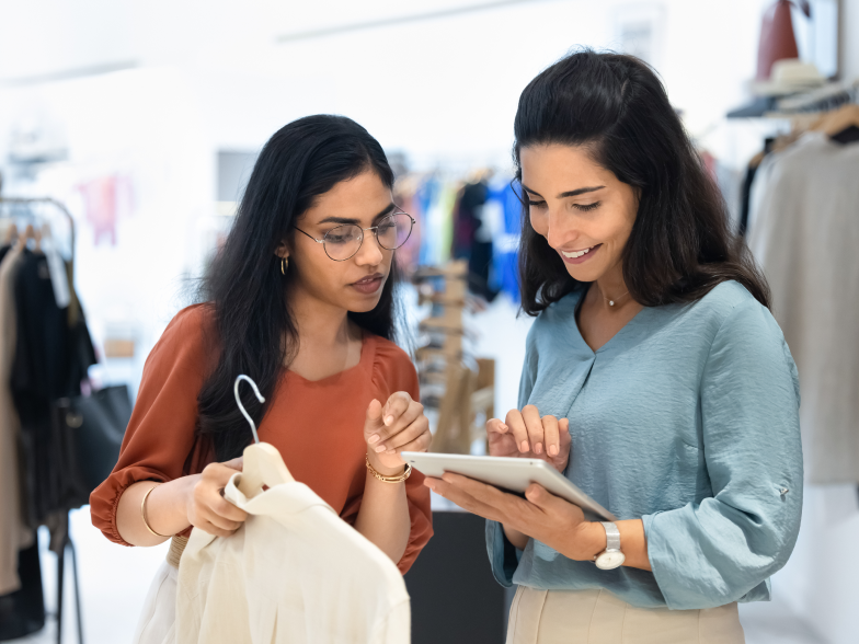 Retail associate checks a customer’s details on a tablet while helping a smiling shopper who’s holding a white blouse in a clothing store