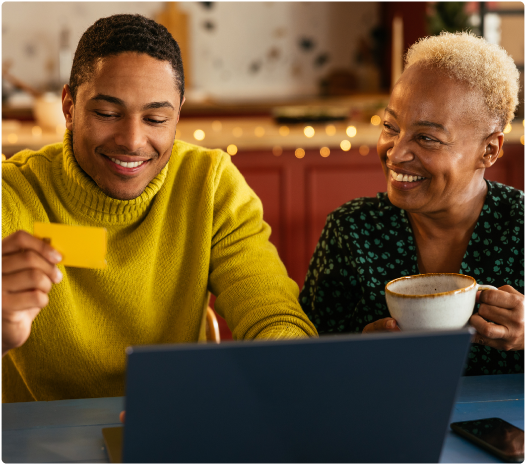 A smiling young man in a yellow sweater holds a card while looking at a laptop. Beside him, an older woman with short hair holds a coffee mug, also smiling warmly. Cozy and cheerful atmosphere.