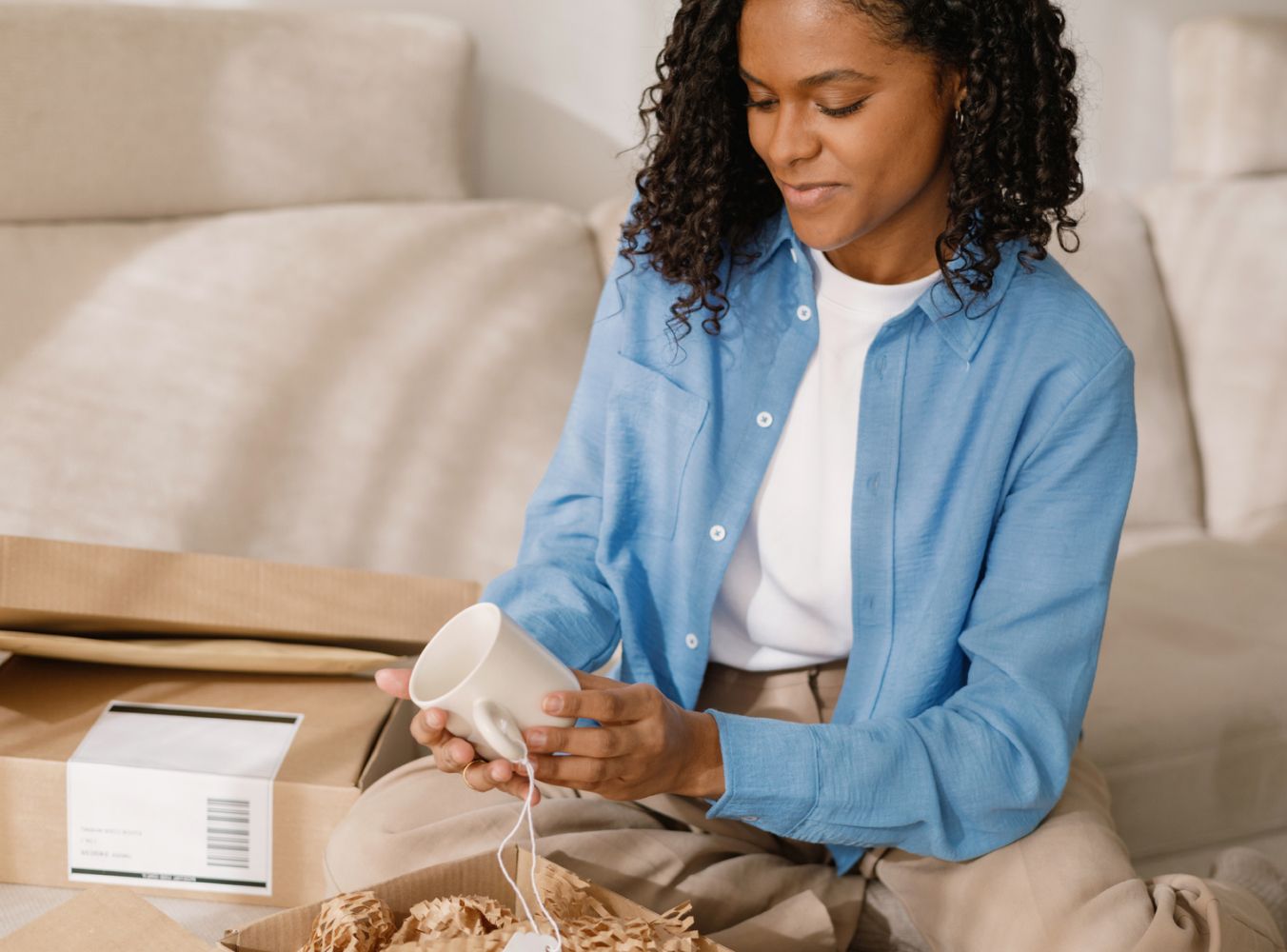 A woman in a blue shirt smiles while unboxing a white mug. She sits on a beige couch with unpacked boxes around, conveying a sense of delight and anticipation.